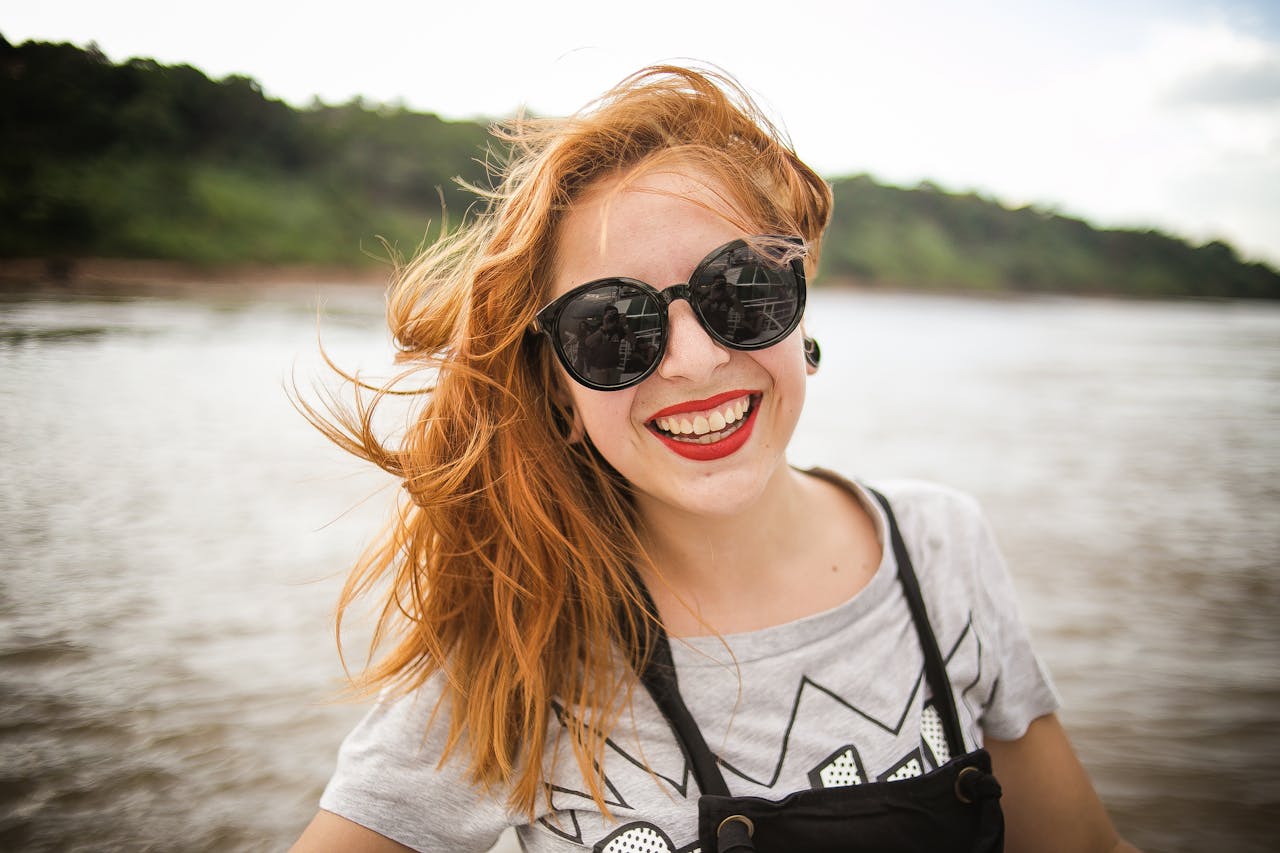 Perte d’autonomie à domicile : que faire concrètement dans les 30 premiers jours ? Smiling young woman with sunglasses and wind-swept hair enjoying a sunny day by the lake.