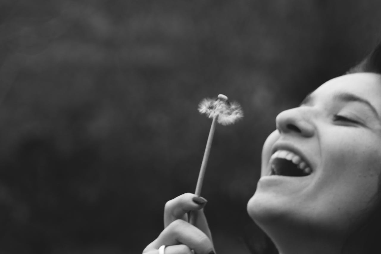 À qui s’adresser concrètement selon votre situation d’autonomie ? A joyful woman smiling while blowing on a dandelion in a black and white portrait.